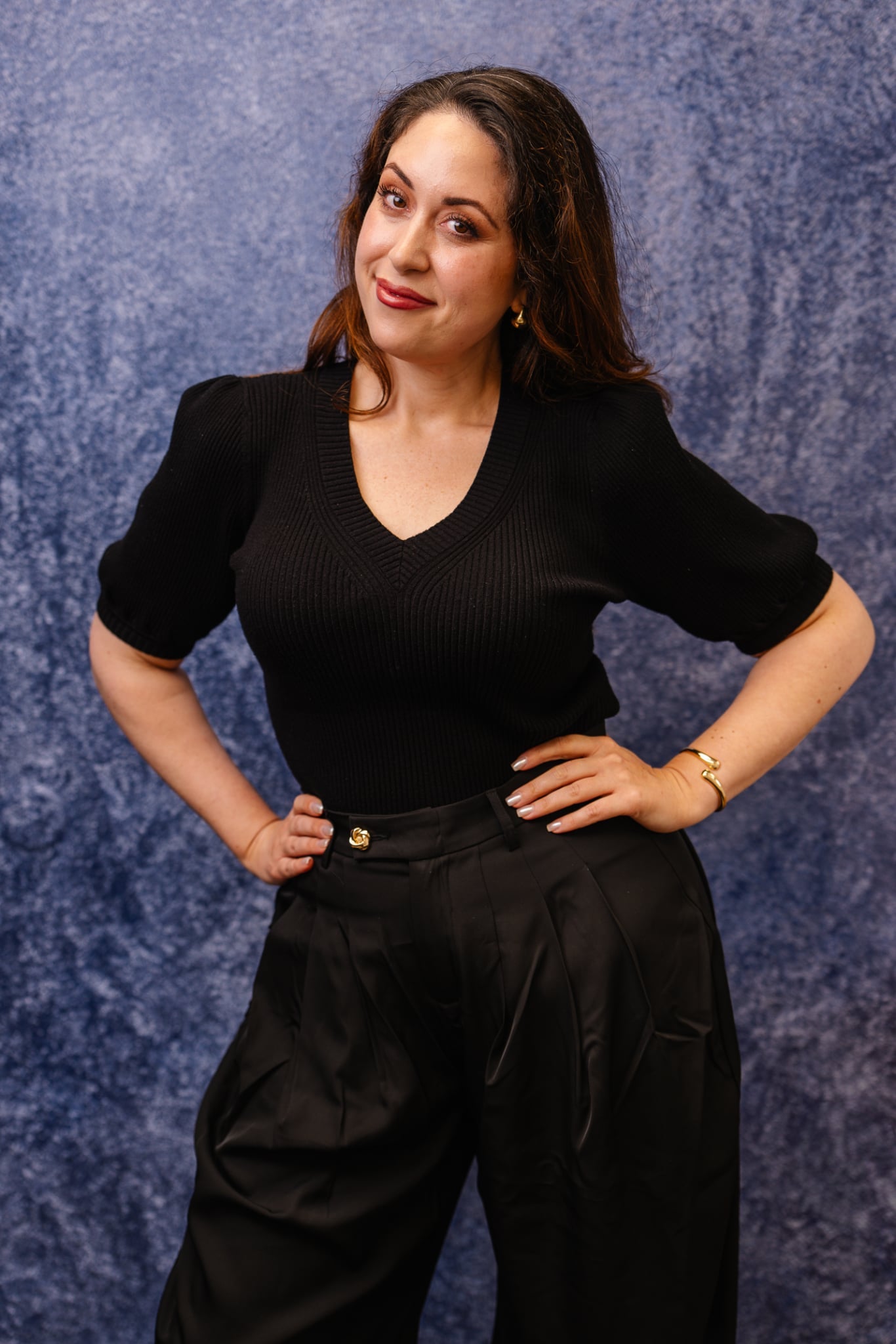 A woman posing in a black short-sleeve ribbed V-neck top and black high-waisted trousers against a textured blue studio backdrop.