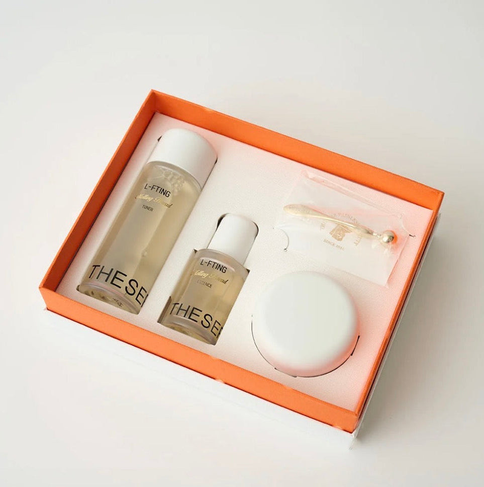 A skincare gift set by THESS from their Lotus Collection, presented in an orange box with a white insert. The set includes a large clear bottle of toner, a smaller clear bottle of essence, a white round jar, and a small clear pouch containing a gold-toned hair tool, all on a light gray background.
